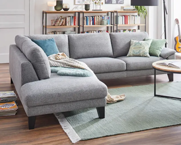 Graues Ecksofa mit Ottomane in modernem Wohnzimmer mit Teppich, Couchtisch und Kissen. Stilvolle Wohnlandschaft für entspanntes Wohnen mit viel Sitzkomfort und zeitlosem Design.